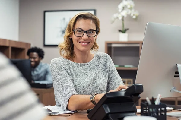 創造的なオフィスで働いていると カメラを見て美しい熟女 コワーキング スペースにコンピューターで作業して受付の肖像画 ヘルプ デスクに座っていると笑顔の眼鏡で成功した実業家 — ストック写真