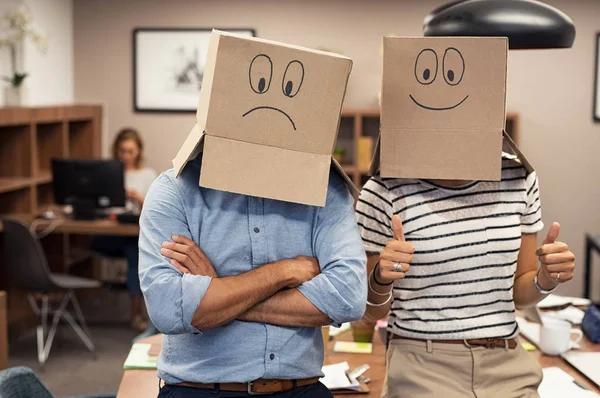 Business man and businesswoman covering face with cardboard box showing happy and sad face. Businessman with folded arms wearing sad face near his colleague with positive expression. Young woman with thumbs up gesture sign at office.