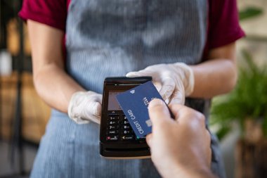 Garson, kredi kartı okuyucu makinesini tutuyor ve tezgahta tek kullanımlık eldiven takıyor. Müşteri kredi kartı tutuyor. Faturayı kredi kartıyla ödeyen adam. Müşteri eli NFC teknolojisiyle irtibatsız kredi kartıyla ödeme yapıyor.. 