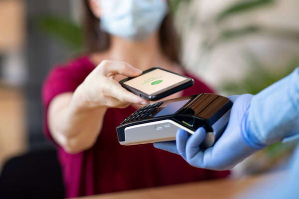 Close up hand of customer paying with smartphone. Cashier hand holding credit card reader machine and wearing protective disposable gloves at bar counter, while client holding phone for NFC payment. Woman wearing face mask while paying bill with mobi