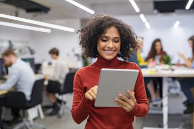 Ofiste dijital tablet kullanan kıvırcık saçlı mutlu iş kadını. Dizüstü bilgisayarda çalışan başarılı genç bir kadın. İş yerinde arka planda oturan meslektaşlarıyla sıradan bir melez iş kadını gibi gülümsüyor..