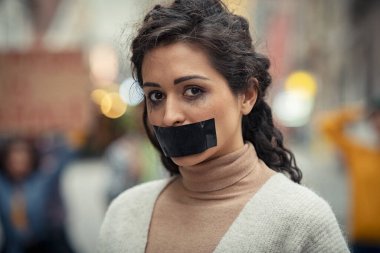 Portrait of young latin woman with black tape on mouth fighting against domestic violence. Abused girl tired of harassment looking at camera. Speechless indian woman with makeup running in female abusive violence campaign on street. clipart