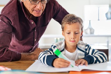Yaşlı adam, çocuğun ödevini evde bitirmesine yardım ediyor. Odaklanmış okul çocuğu, büyükanne ve büyükbabasıyla deftere proje hazırlıyor. Torun ve büyükbaba kağıt üzerinde yazma alıştırması yapıyorlar..