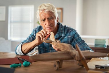 Focused senior man using sandpaper to polish wooden airplane sculpture. Mature carpenter working on making wooden flight at home. Happy old grandfather at home busy making handmade sculpture. clipart