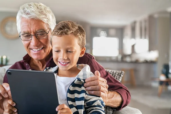 快乐的老人带着孩子在家里用数字平板电脑进行视频通话 笑爷爷和孙子在家里玩数码平板电脑 快乐的小男孩坐在爷爷的膝上 在笔记本电脑上看带有复制空间的卡通片 — 图库照片