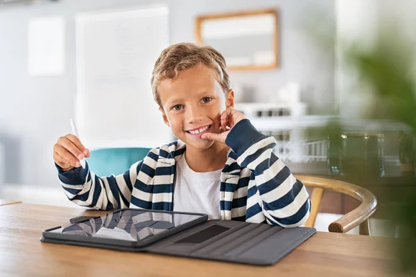 Child drawing picture with digital pen on tablet while looking at camera. Portrait of smiling cute kid using stylus on tablet to do homework for school. Artistic boy drawing on digital tablet using stylus at home and looking at camera.