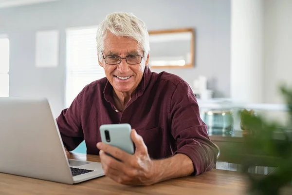 在笔记本电脑前使用智能手机的成熟男人 快乐的老生意人一边在家工作一边用手机发短信 快乐微笑的老人使用社交媒体网络技术 感到兴奋 — 图库照片