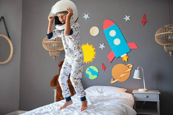 可爱的小男孩穿着宇航员的服装 背着火箭和行星在床上跳着 戴着宇航员头盔的孩子在玩耍 梦想着成为一名宇航员 穿着太空睡衣和与太阳系和行星一起玩的快乐的孩子D — 图库照片