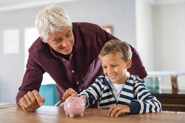 开心地笑着孙子和爷爷一起在储蓄罐里加钱 年长的男人和可爱的男孩在家里把硬币放进粉红色的储蓄罐 兴奋的男孩在爷爷的帮助下存钱 — 图库照片