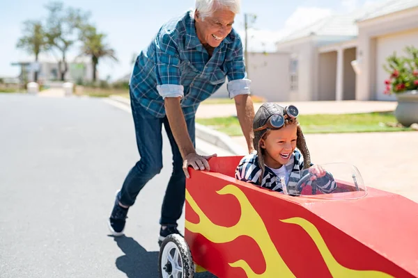 おじいさんは孫が子供のおもちゃの車に乗るのを手伝っている 男の子が通りを走っている間 幸せな老人は車を押している 祖父母はゴーカートで男の子と遊ぶ — ストック写真