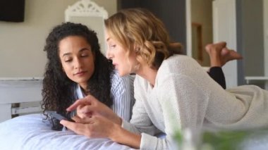 Yatakta yatıp akıllı telefon kullanarak online alışveriş yapan neşeli çok ırklı bayan arkadaşlar. İki genç kadın cep telefonundan komik videolar izleyip birlikte gülüyorlar. Lezbiyen çift yatakta dinlenirken cep telefonu kullanıyor..