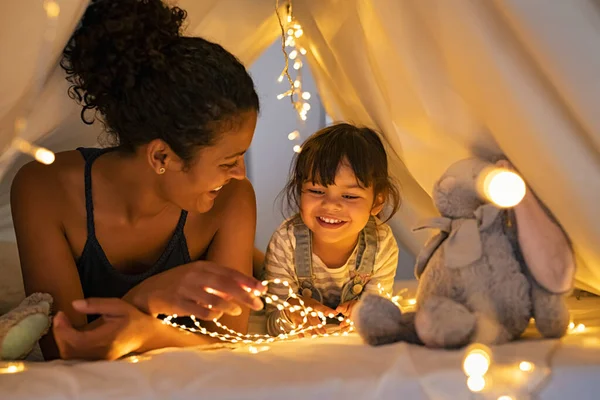 遊び心のある母親と娘は 明るいテントの中で子供部屋で遊んでいます アフリカのお母さんと素敵なかわいい女の子は 自宅で夜に子供小屋で楽しんでいます 母と娘はストリップライトと一緒に時間を過ごす — ストック写真