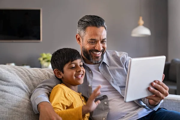 インド人の父と笑顔の息子は自宅でデジタルタブレットを使用してソファに座っている 男と男の子は一緒に時間を過ごしながら自宅でビデオ通話のためのデジタルタブレットを使用しています 隔離中にビデオ通話を行う息子と中東のお父さん — ストック写真