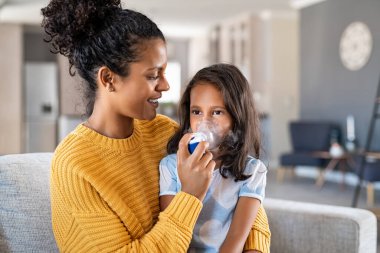 Little indian girl making inhalation with nebulizer at home with lovely mother. Woman makes inhalation to a sick child while embracing her. Mom helping daughter with cold and flu to inahale nebuliser aerosol sitting on couch. clipart