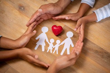 Aile kâğıdını kırmızı kalple koruyan baba, anne ve kızların ellerini kapatın. Kağıt kesiği ve ahşap arka planda kırmızı kalp şeklinde bir taş olan melez bir ailenin elleri. Birlik, sigorta ve aşk kavramı.