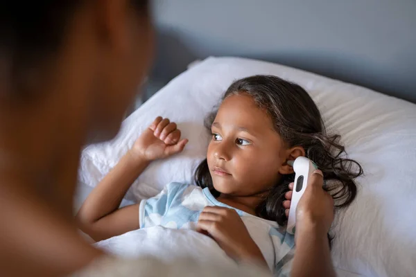 小さな病気の女の子の温度を確認するために耳の温度計を使用してアフリカのお母さんの閉じるベッドの上に横たわる インド人の娘は熱をチェックしながらベッドで休んでいます 風邪やインフルエンザの症状を持つ少女の肖像画一方 母親が隣に座って聞く — ストック写真