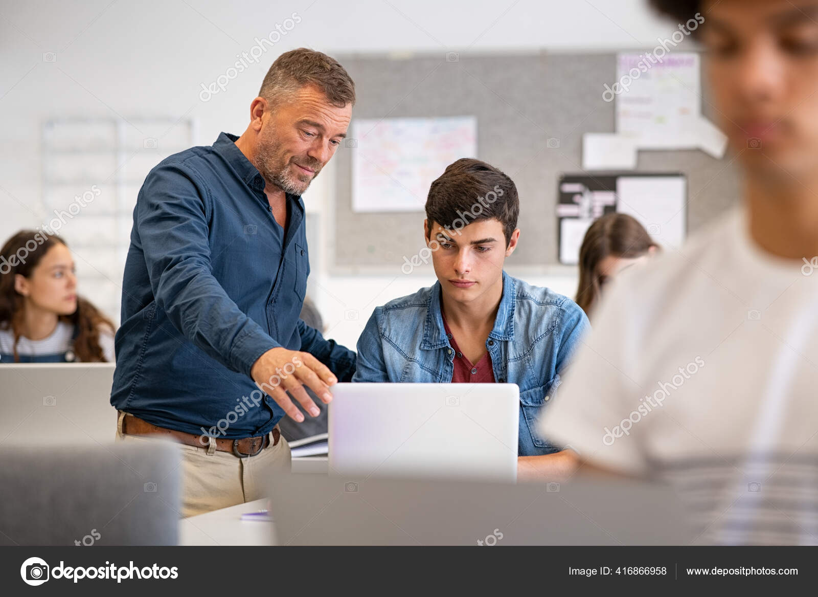 教授は コンピュータレッスン中に教室でラップトップで大学生を支援 先生は机に座っている男に何かを話して説明します 大学での講義中にラップトップを持つ 高校生の若者を助ける熟女講師 ストック写真 C Ridofranz
