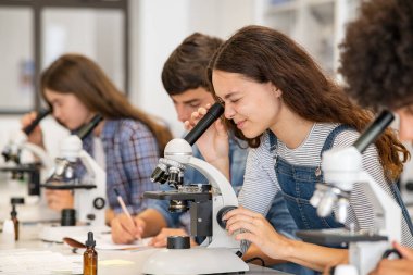 Çok ırklı üniversite öğrencileri bilim laboratuarında mikroskop kullanarak deney yapıyorlar. Üniversite ogrencisi masasinda siraya otururken biyoloji sinifinda mikroskopla bakiyor. Liseli kız ders sırasında örnekleri inceler.