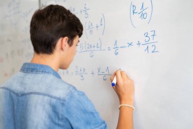 Lise öğrencisinin matematik problemini sınıfın beyaz tahtasında çözdüğü arka plan. Kalem kullanarak beyaz tahtaya matematik çözeltisi yazan genç adam. Ders sırasında matematik ifadesini çözen üniversiteli çocuk..