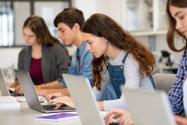 Kütüphanede dizüstü bilgisayarda oturan ciddi üniversite öğrencileri. Genç üniversite çok ırklı öğrencileri sınıfta çalışmak için bilgisayar kullanıyor. Bilgisayar dersi sırasında sıradan bir kızın dizüstü bilgisayarda yazışının yan görüntüsü.
