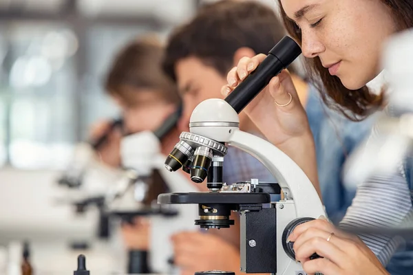 Close up of young woman seeing through microscope in science laboratory with other students. Focused college student using microscope in the chemistry lab during biology lesson. University students studying in high school science laboratory.