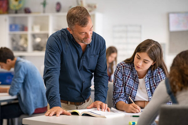 Secondary student being helped by professor during class. Mature man lecturer helping young woman during class test. High school girl in a lecture asking for explanations to helpful teacher. 