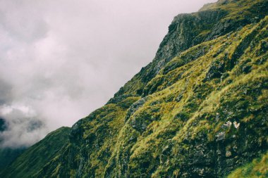 Bulutların arkasında yeşil bir kayalık dağ.