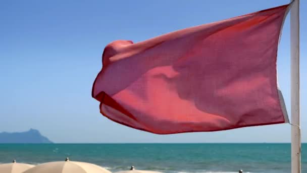 rote Fahne weht im Wind am Strand in der Nähe weißer Sonnenschirme mit einem Berg am Horizont Landschaft an einem blauen Himmel sonnigen Sommertag. keine Menschen.