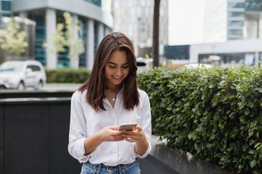 Mutlu işkadını cep telefonuyla ofisi, açık havada yürüyüş gülümseyen güzel kadın tarama telefon, kadın Yöneticisi manifatura yakınındaki modern smartphone kullanarak bir messenger uygulaması 