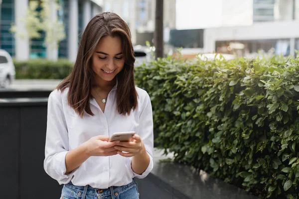 Mutlu işkadını cep telefonuyla office, güzel kadın telefon gülümseyen açık havada yürüyüş tarama, modern smartphone kullanarak kadın Yöneticisi manifatura yakınındaki. Kopya alanı metin veya tasarım için