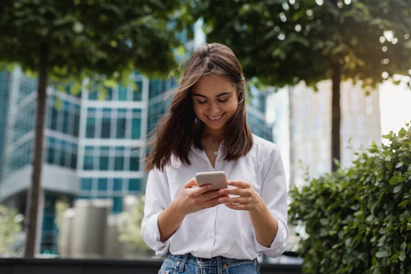 Mutlu işkadını cep telefonuyla ofisi, açık havada yürüyüş gülümseyen güzel kadın tarama telefon, kadın Yöneticisi manifatura yakınındaki modern smartphone kullanarak bir messenger uygulaması 