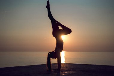 Gün batımında sahilde güzel bir poz elinde duran genç jimnastikçi silüeti. Güneş geliyor up ve kadının iskelede egzersiz yapması. Açık havada handstanding pratik profesyonel yoga öğretmeni