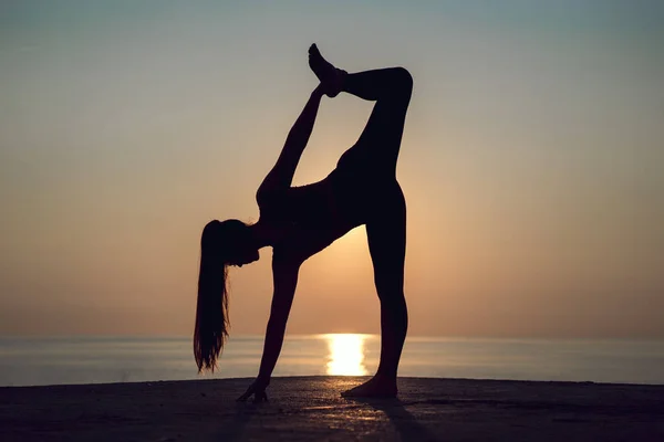 Silhouette of young woman practicing yoga alone on the beach at sunset. Female happiness, fitness and healthy lifestyle. Sports girl doing stretching and exercise outdoors during vacation