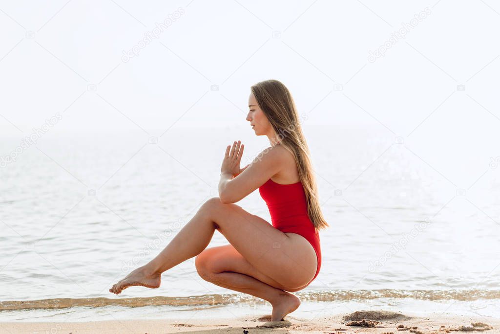 Young woman practice balance asanas on summer yoga session on a beautiful golden beach of Spain on a yoga tour. Girl in red swimsuit meditating on the beach and doing yoga or pilates to be healthy