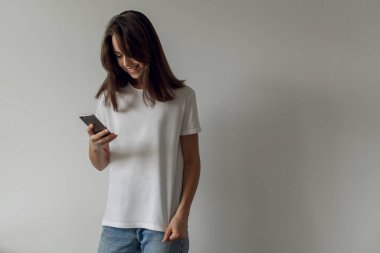 Fotoğraf üzerinde bir beyaz duvar arka plan evde duran bir hareket eden smartphone kullanarak geçici bir beyaz t-shirt giyen bir gülümseyen mutlu hippi kız. Genç kadın cep telefonu ile yazarak