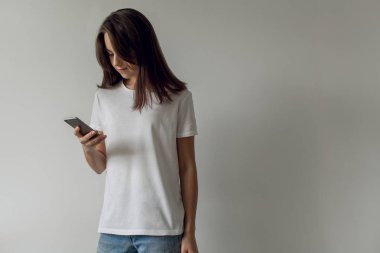 Hippi erkek elinde beyaz bir duvar arka plan üzerinde modern smartphone ayakta tutan. Boş t-shirt logo, metin veya tasarım için boş yer ile. Online alışveriş kavramı.