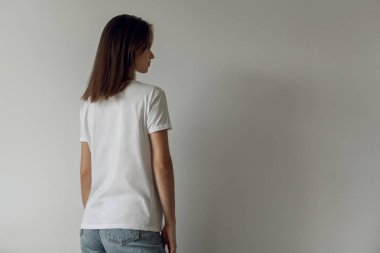 Arka planda, arkadan görünüm mavi kot pantolon ve logo veya tasarım, beyaz tişört, beyaz duvar mock-up için alanı boş beyaz tişört giyen genç hippi kız