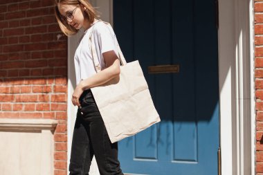 Genç hipster gülümseyen kız beyaz boş pamuk alışverişçi kağıt torba giymiş beyaz tişört ve siyah kot, mock-up boş çanta, tuğla duvar ve mavi kapı arka planda, alışveriş kavramı ile