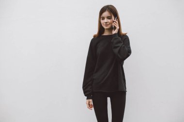 Genç hipster gülümseyen kız giyiyor boş siyah hoodie ve kot pantolon ve cep telefonu, boş siyah kazak, mock-up konuşurken arka plan beyaz