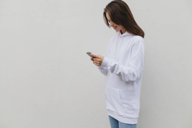 Bir beyaz beton duvar arka plan üzerinde duran bir hareket eden smartphone kullanarak geçici bir beyaz kazak giyen bir gülümseyen mutlu hippi kız fotoğrafı