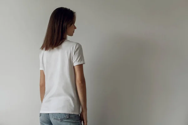 Arka planda, arkadan görünüm mavi kot pantolon ve logo veya tasarım, beyaz tişört, beyaz duvar mock-up için alanı boş beyaz tişört giyen genç hippi kız