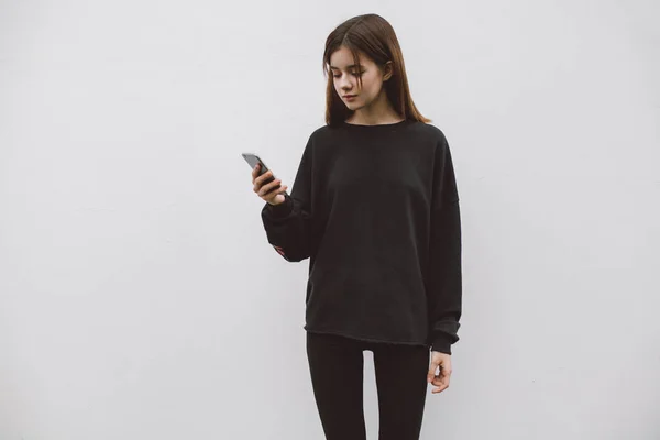 Hippi erkek elinde beyaz bir duvar arka plan üzerinde modern smartphone ayakta tutan. Boş hoodie logosu, metin veya tasarım için boş yer ile. Siyah boş kazak mockup
