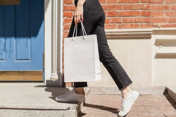 Genç hipster gülümseyen kız beyaz boş kağıt torba giymiş beyaz tişört ve siyah kot, mock-up boş çanta, tuğla duvar ve mavi kapı arka planda, alışveriş kavramı ile