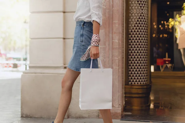 Attractive woman doing shopping. Happy girl with blank white shopping bag walking on city streets and shops. Consumerism, shopping, lifestyle concept. Copy space for your logo