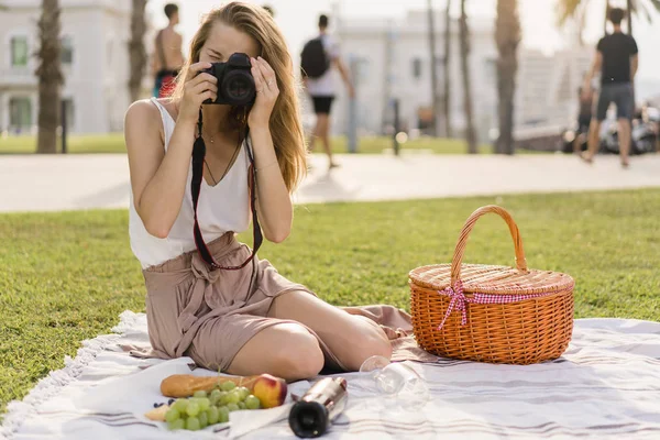 Genç güzel kız bir kamu park açık havada piknik ve şarap ve meyve ile bir öğle yemeği sırasında bir makinesinde anılar için erkek arkadaşı resim çekmek. Kadın pembe etek fotoğraf makinesi kullanma