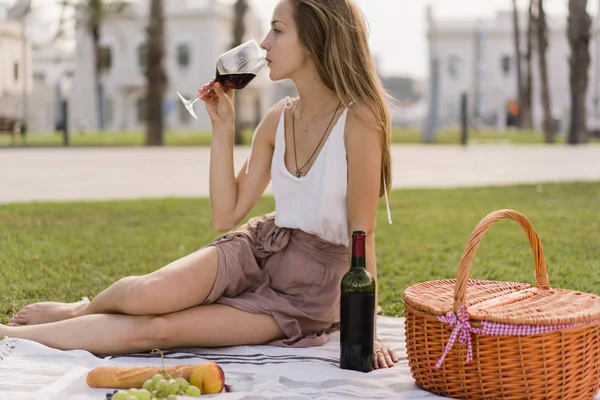 Pretty woman having picnic in the park, sitting on a green grass with a blank bottle of red wine and snacks, enjoying holidays and summer, mockup of a bottle of wine with a space for your logo