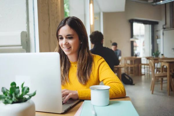 Çekici olumlu genç kadın, kahve dükkanında yüksek hızlı 4G kablosuz internete bağlı modern laptop cihazıyla teknoloji haberlerini kontrol ediyor.
