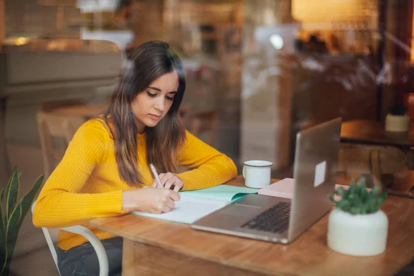 Kafe kızında modern dizüstü bilgisayarla çalışan güzel genç bir öğrenci çevrimiçi okulda 4G internete bağlı bir kafede notlar alıyor.