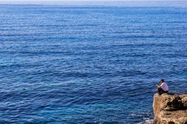 Okyanus kıyısında, arkasında mavi su ve gökyüzü olan, taşın üzerinde tanımlanamayan kıdemli bir balıkçı. 