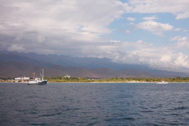 Beyaz gemi kıyıdan ayrılıyor. Kırgızistan Dağları. Okyanus denizindeki mavi su. Gökyüzünde güzel bulutlar. Adanın denizine git. Denizde tatil. Hava turizmi. Yüksek kalite fotoğraf